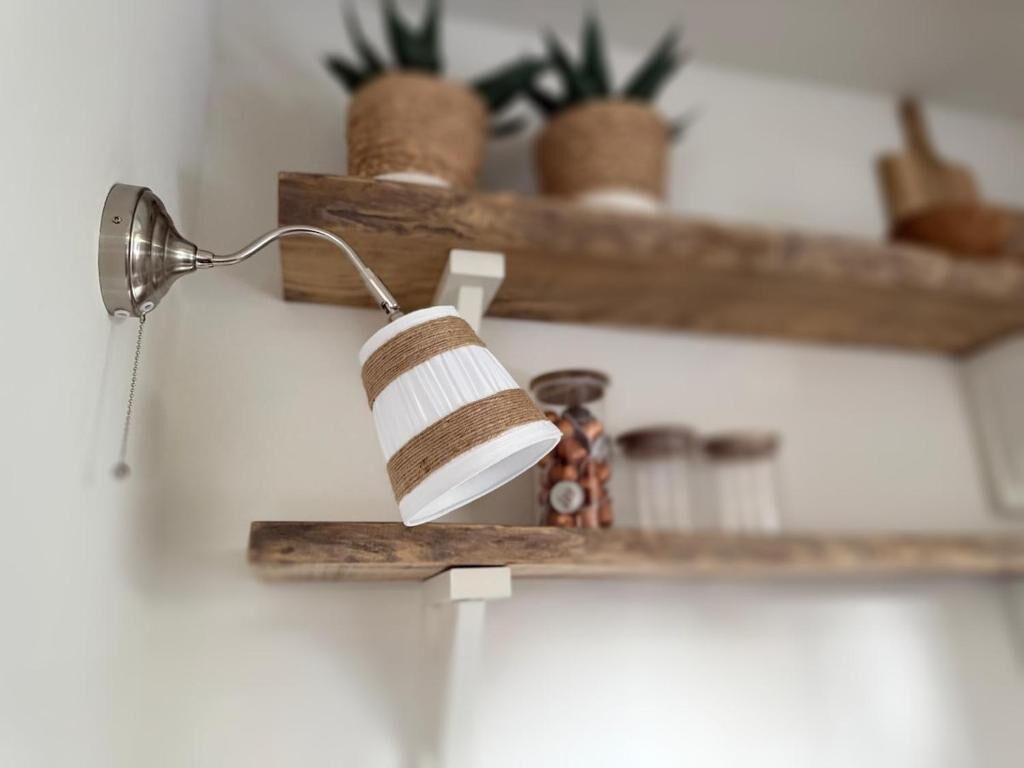 Una lámpara rústica de pared con diseño de cesta tejida, estantes de madera con plantas en macetas y frascos.