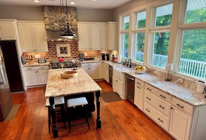 A kitchen with white cabinets, a granite countertop, a large island with stools, a stone backsplash, and a view through large windows.