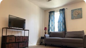 Uma sala de estar com televisão de tela plana em um suporte, sofá, armário, luminária e janela com cortinas.