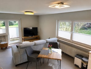 A modern living room with a sectional sofa, a television on a wooden stand, a dining table with chairs, and large windows with blinds. - Vacation with the Winemaker, Use of the Garden—Private Restaurant (Burgen)