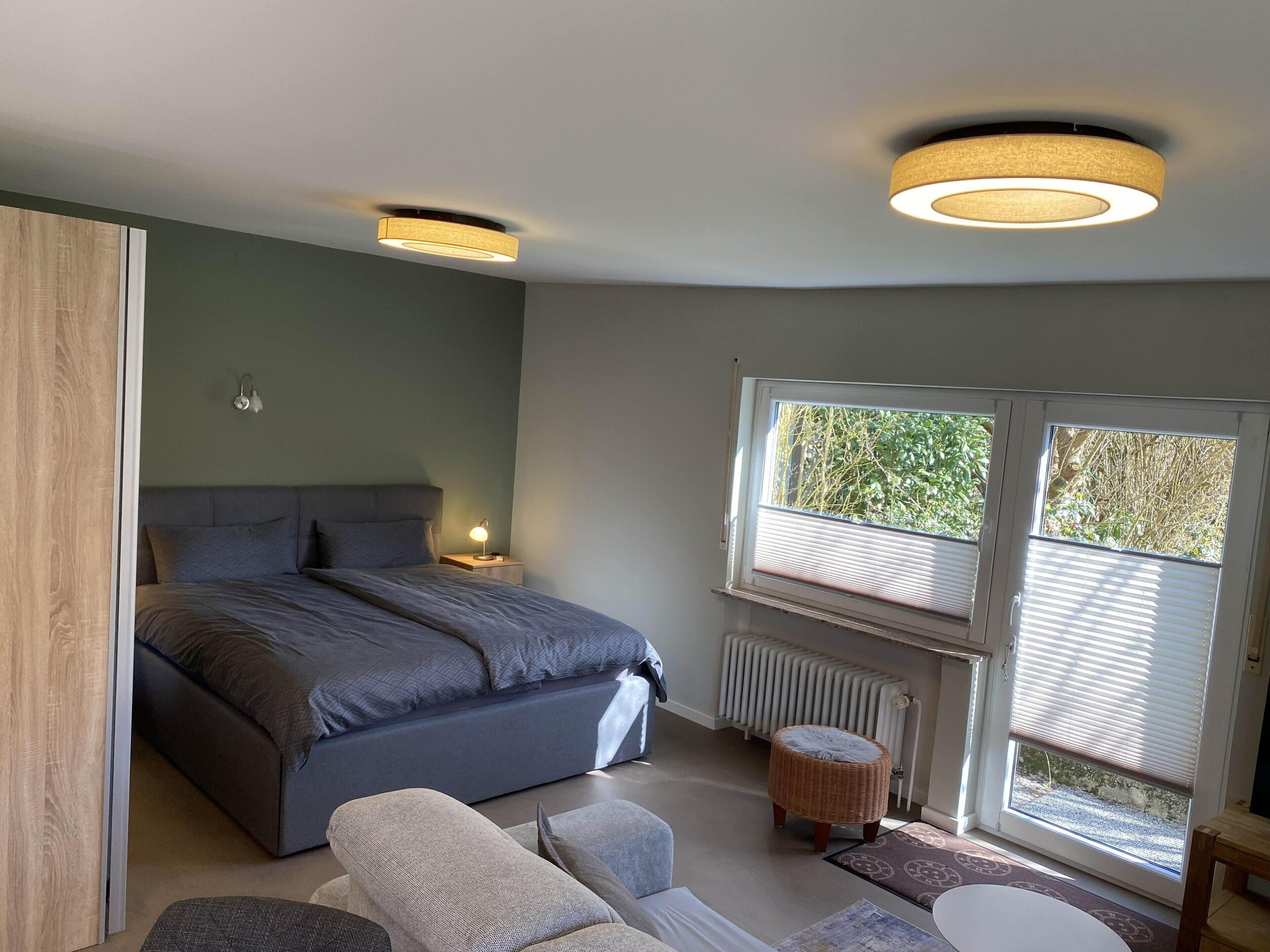 Une chambre à coucher avec un lit, des lampes de chevet, une fenêtre avec des stores, un radiateur et un tabouret en osier.