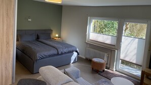 Une chambre à coucher avec un lit, des lampes de chevet, une fenêtre avec des stores, un radiateur et un tabouret en osier.