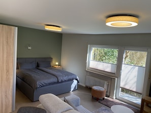 Un dormitorio con una cama, lámparas de noche, una ventana con persianas, un radiador y un taburete de mimbre.