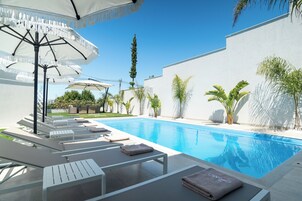 A poolside area with lounge chairs and umbrellas, a clear blue pool, and a white wall.