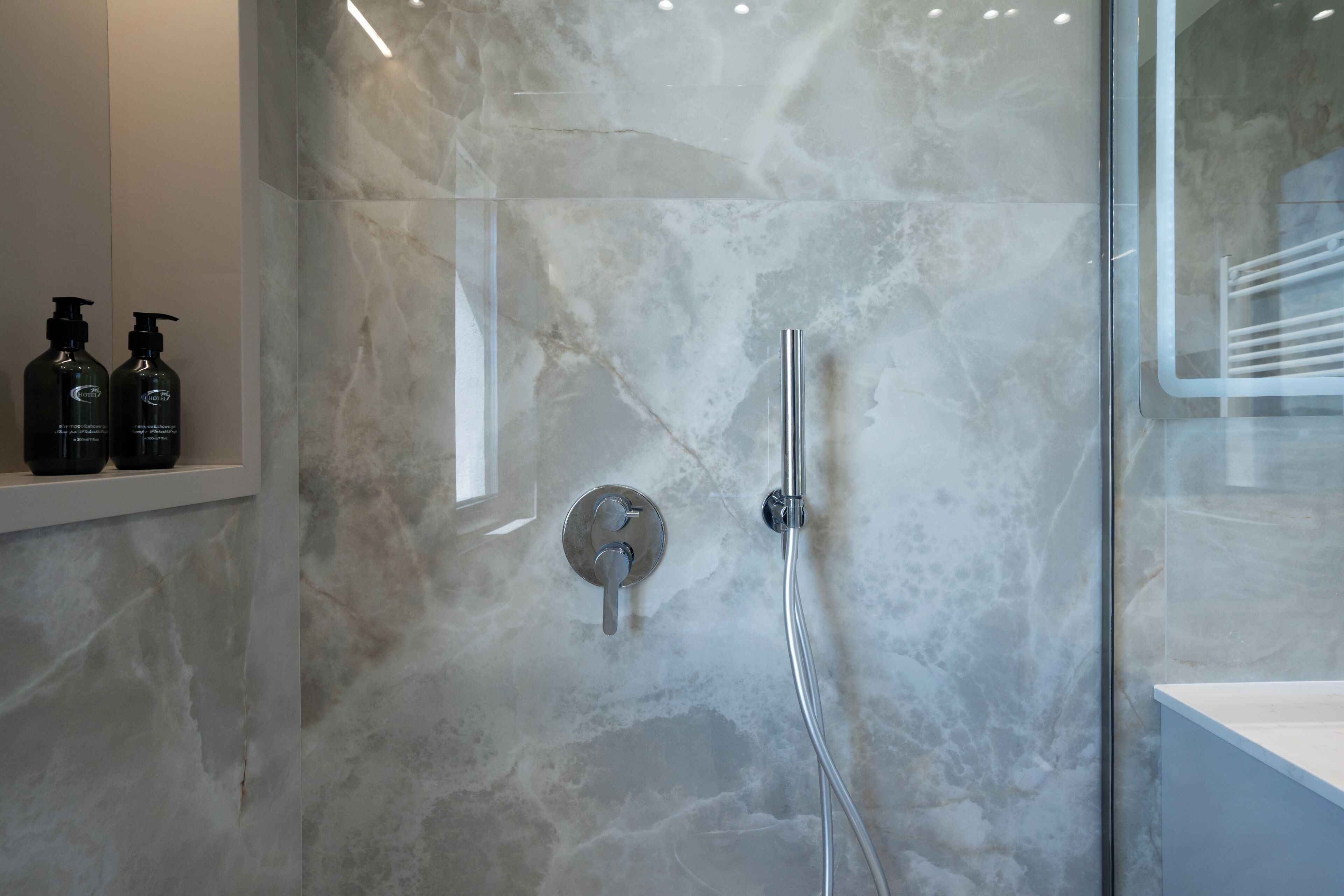 Ein modernes Badezimmer mit einer Glastrennwand-Dusche, Marmorwänden und einem Ablagefach mit Pflegeprodukten.