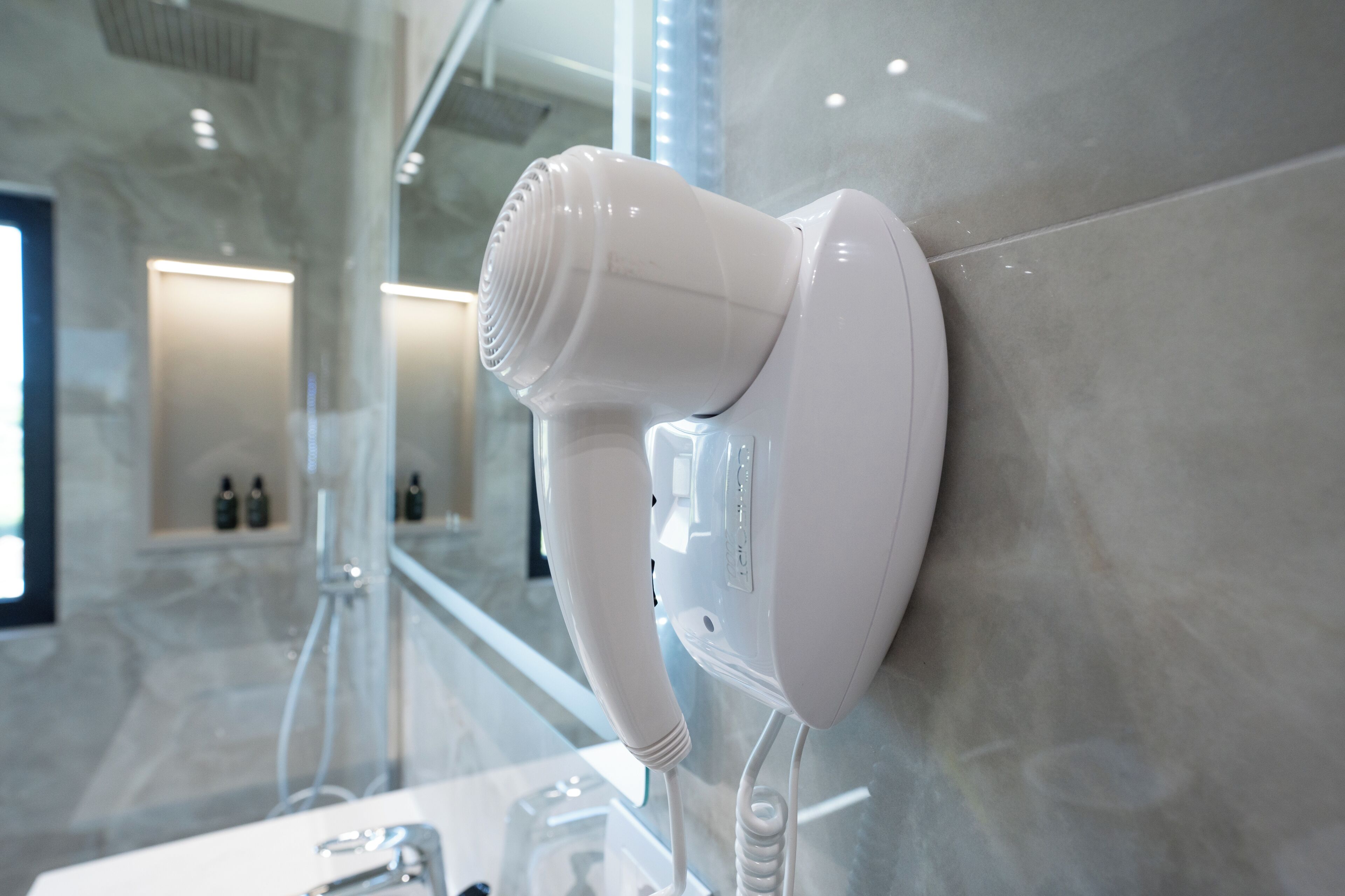 A hair dryer mounted on a wall in a modern bathroom with a glass shower enclosure and a marble floor.