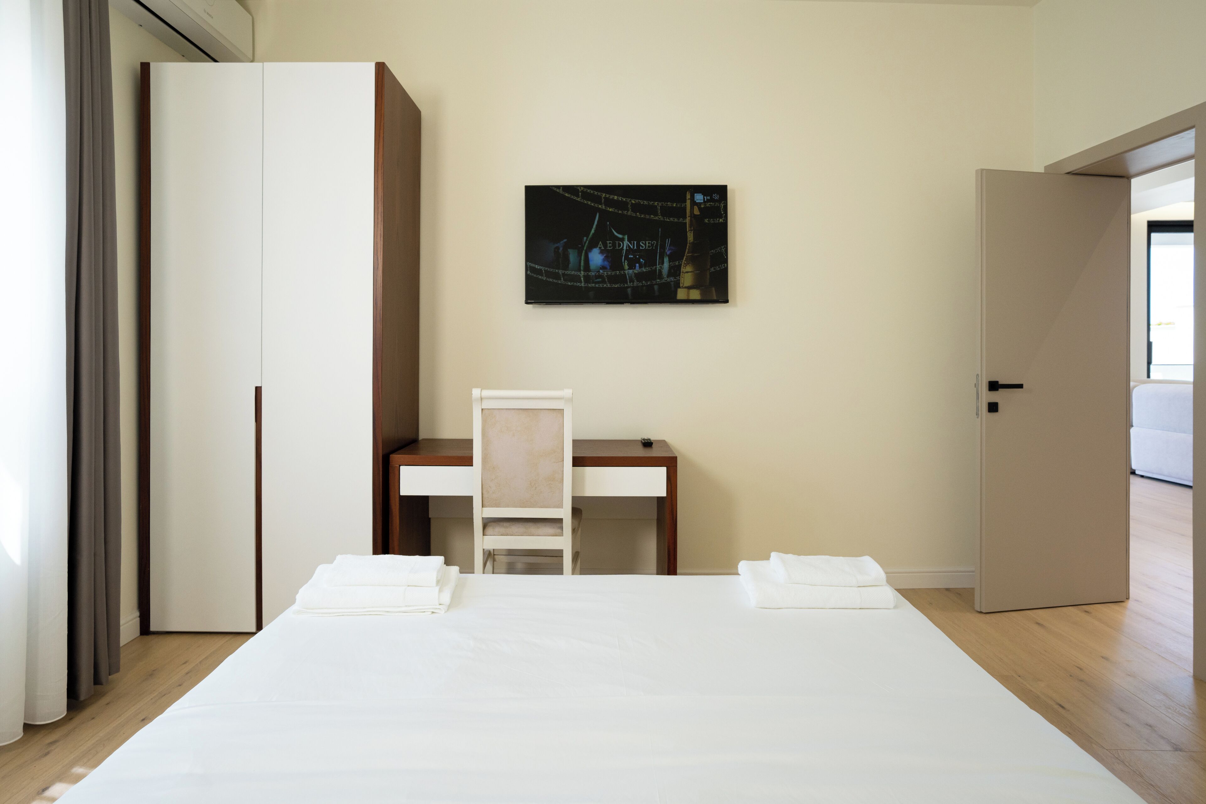 A hotel room with a large bed, a desk, and a chair. There is a painting on the wall and a door to the right.