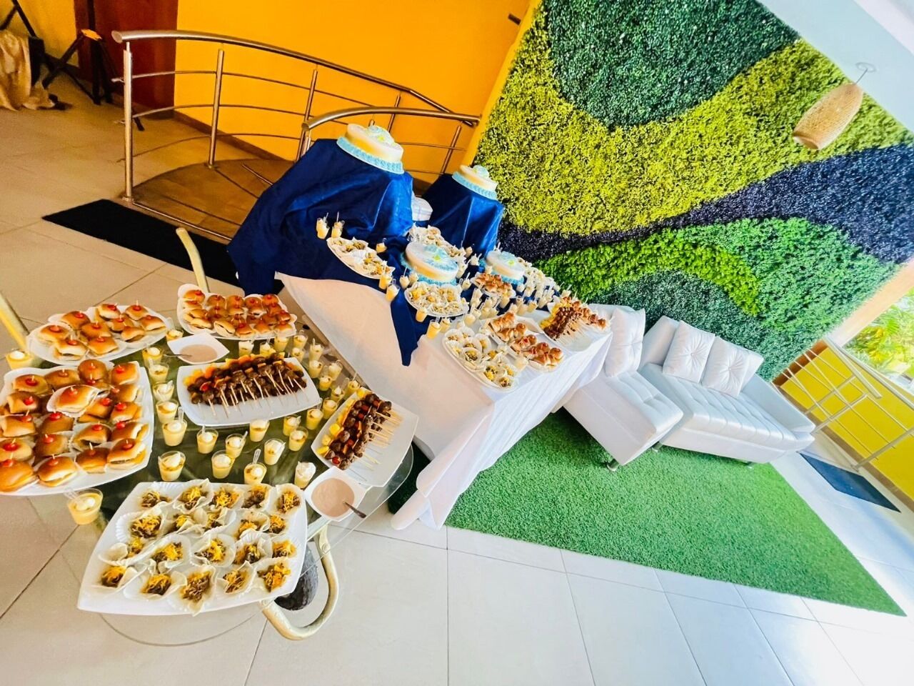 A buffet table with various food items and a white sofa in a room with green artificial grass and a yellow wall.