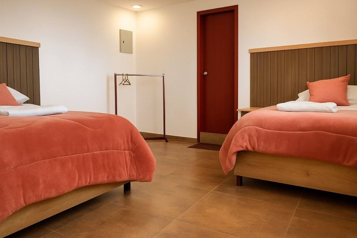 A hotel room with two beds, each with a wooden headboard and orange bedding. There is a wooden floor, a door, and a small table with a lamp.