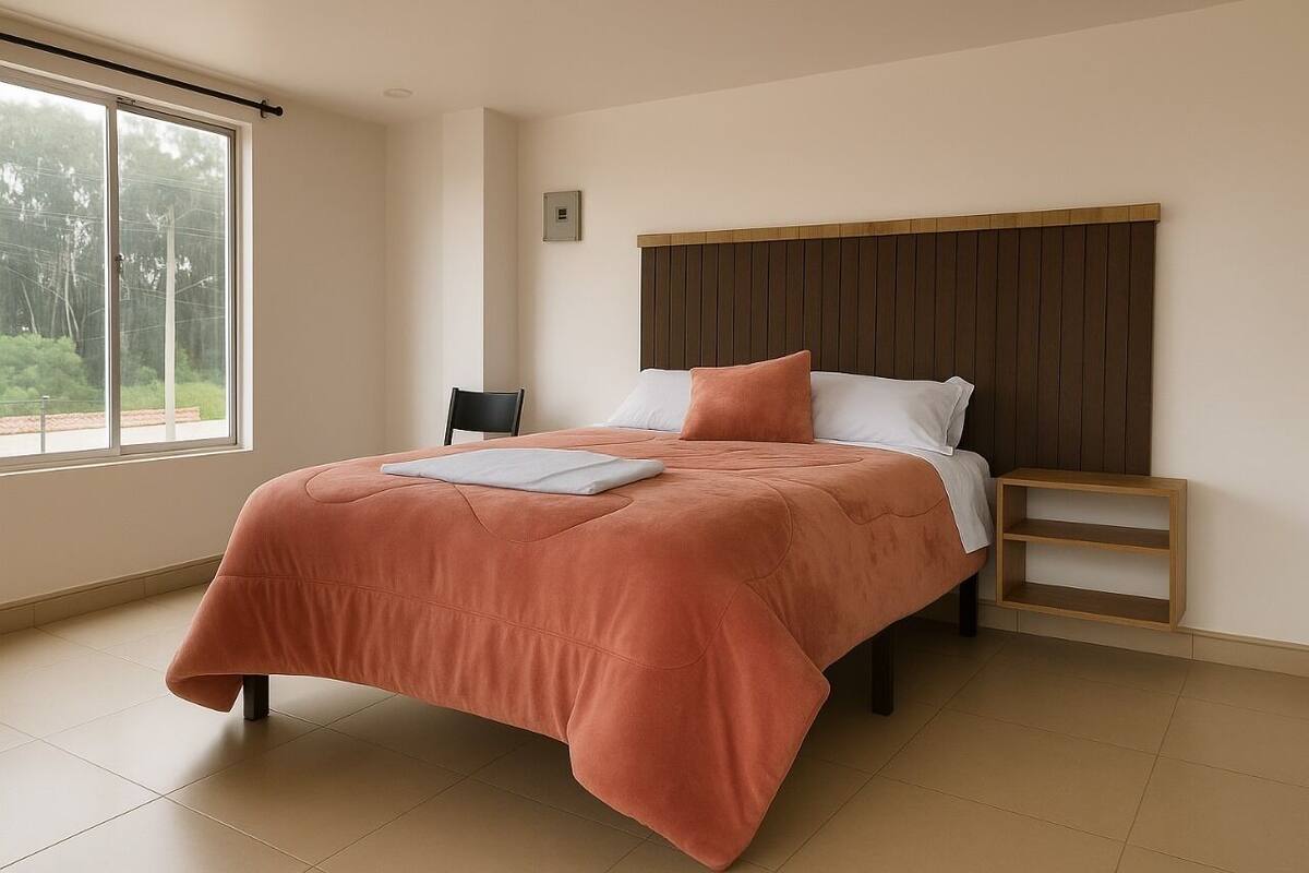 A modern bedroom with a large bed, a wooden headboard, a small bedside table, and a window with a view of greenery.
