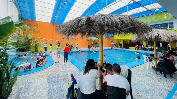 An indoor swimming pool with a thatched roof structure, surrounded by people and a few chairs. - HOSTERIA DON POLY (San Miguel de Salcedo)
