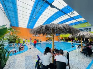 An indoor swimming pool with a thatched roof structure, surrounded by people and a few chairs. - HOSTERIA DON POLY (San Miguel de Salcedo)