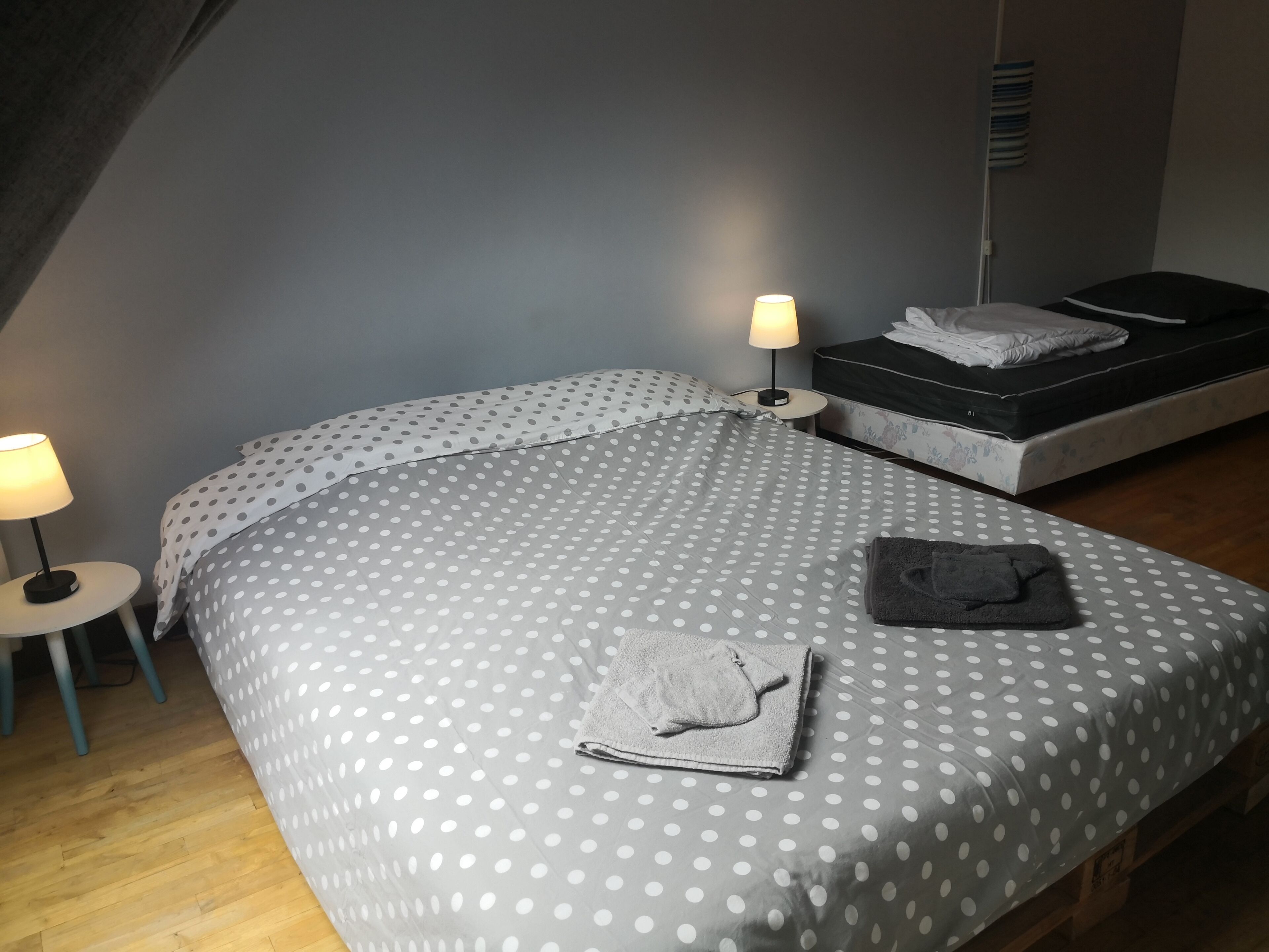 Un dormitorio con una cama cubierta con ropa de cama de lunares, una lámpara de noche, una mesita y un banco con una toalla.
