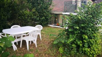 Un jardín con una mesa y sillas blancas, rodeado de abundante vegetación y una casa con tejado de tejas.