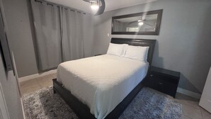 Une chambre à coucher comprenant un lit, une table de chevet, un ventilateur de plafond, une fenêtre avec des rideaux et un miroir fixé au mur.