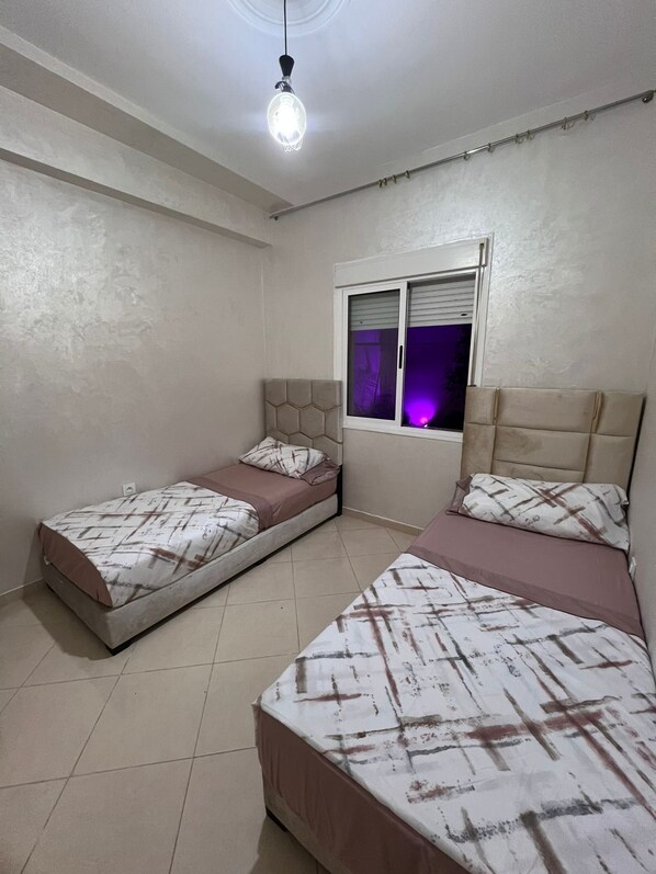 A bedroom with two single beds, a window with blinds, a ceiling light, and a plain wall.