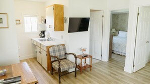 A compact room with a kitchenette, a wooden chair, a small table with a vase, and a bed in the adjacent room. - Gallant Cottage-PEI Charm, updated cottage with AC near Charlottetown. Waterview (Alexandra)