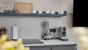 A kitchen with a wooden cabinet, a shelf with white cups and glasses, a paper towel holder, and a coffee machine.