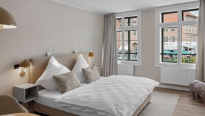 A modern bedroom with a large bed, bedside lamps, a nightstand, a round wooden table, and a view of buildings through a window.