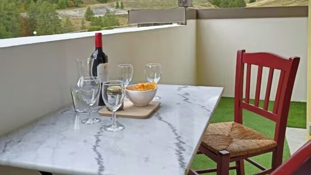 A balcony with a table set for two, a bottle of wine, and a bowl of snacks. The view includes mountains and a valley.