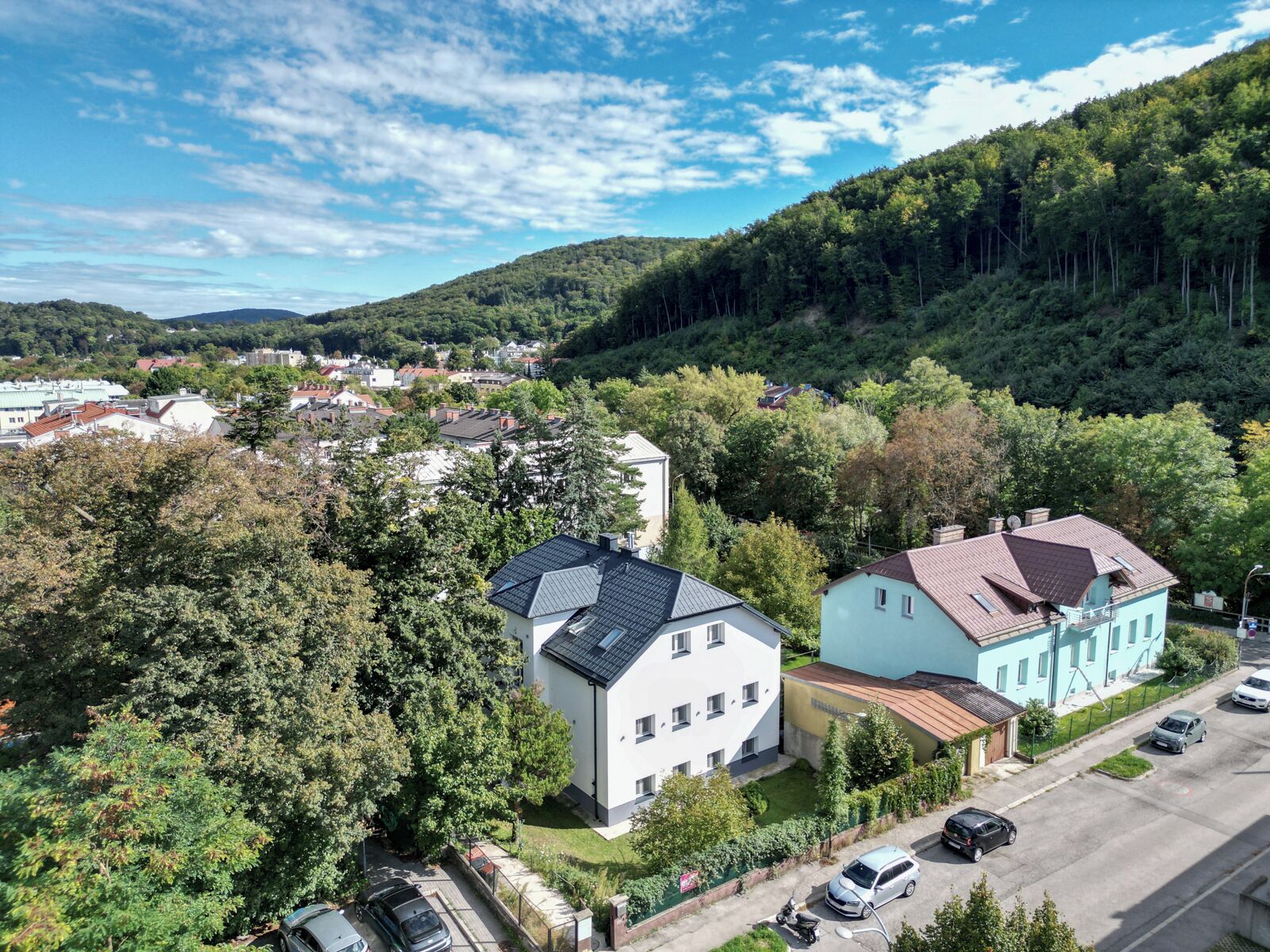 A residential area with houses surrounded by dense greenery and hills in the background.