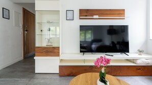 Een moderne woonkamer met een flatscreen televisie aan een witte muur, een houten kastje eronder en een glazen vitrine met decoratieve objecten.