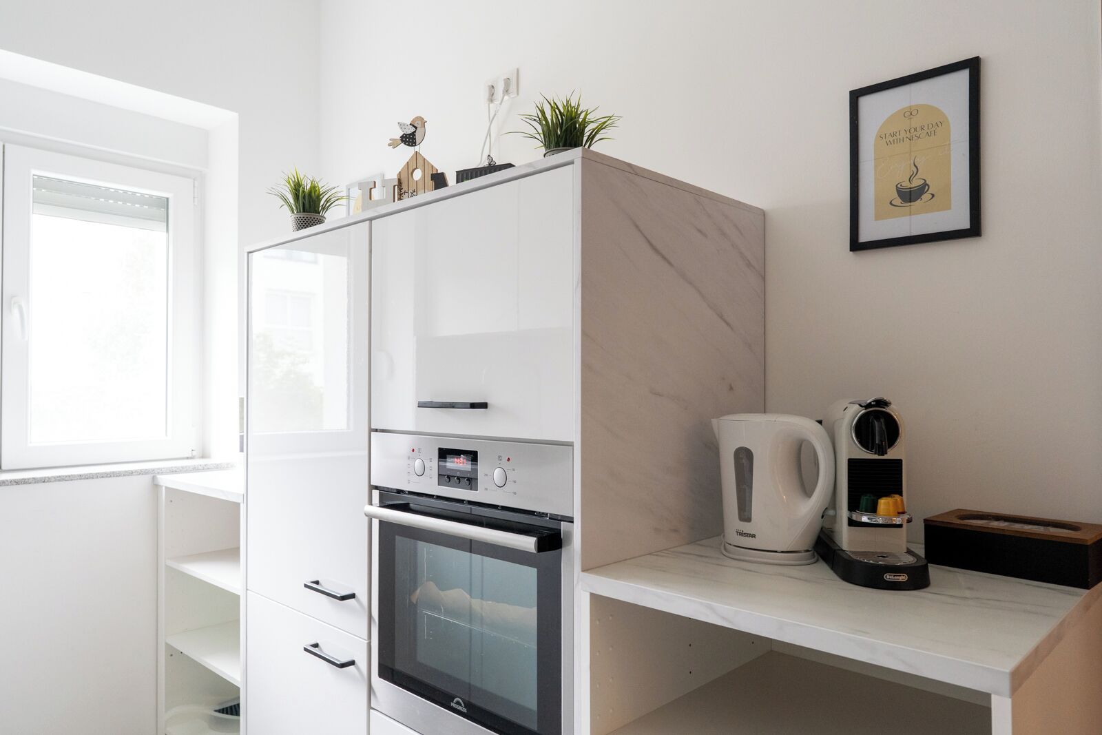 Eine moderne Küche mit weißen Schränken, einem eingebauten Ofen, einer Kaffeemaschine und einem gerahmten Motivationsspruch an der Wand.