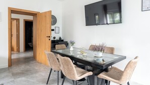 Een moderne eetruimte met een glazen tafel, beklede stoelen, een flatscreen televisie en een houten deur naar een badkamer.