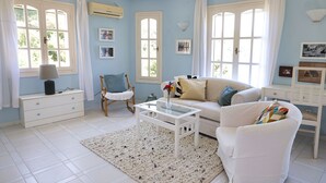 A bright living room with a white sofa, a coffee table, and a chair. There are windows with curtains, a white dresser, and a rug on the floor. - Villa Panorama, the magic of Skiathos in a family villa with history (Skiathos)