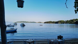 A beachside dining area with tables and chairs, overlooking a calm body of water with several boats and a distant shoreline. - Villa Panorama, the magic of Skiathos in a family villa with history (Skiathos)