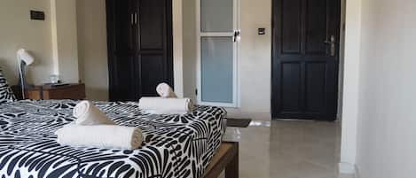 A bedroom with a bed, two dark doors, a bedside table with a lamp, and a rolled towel on the bed.