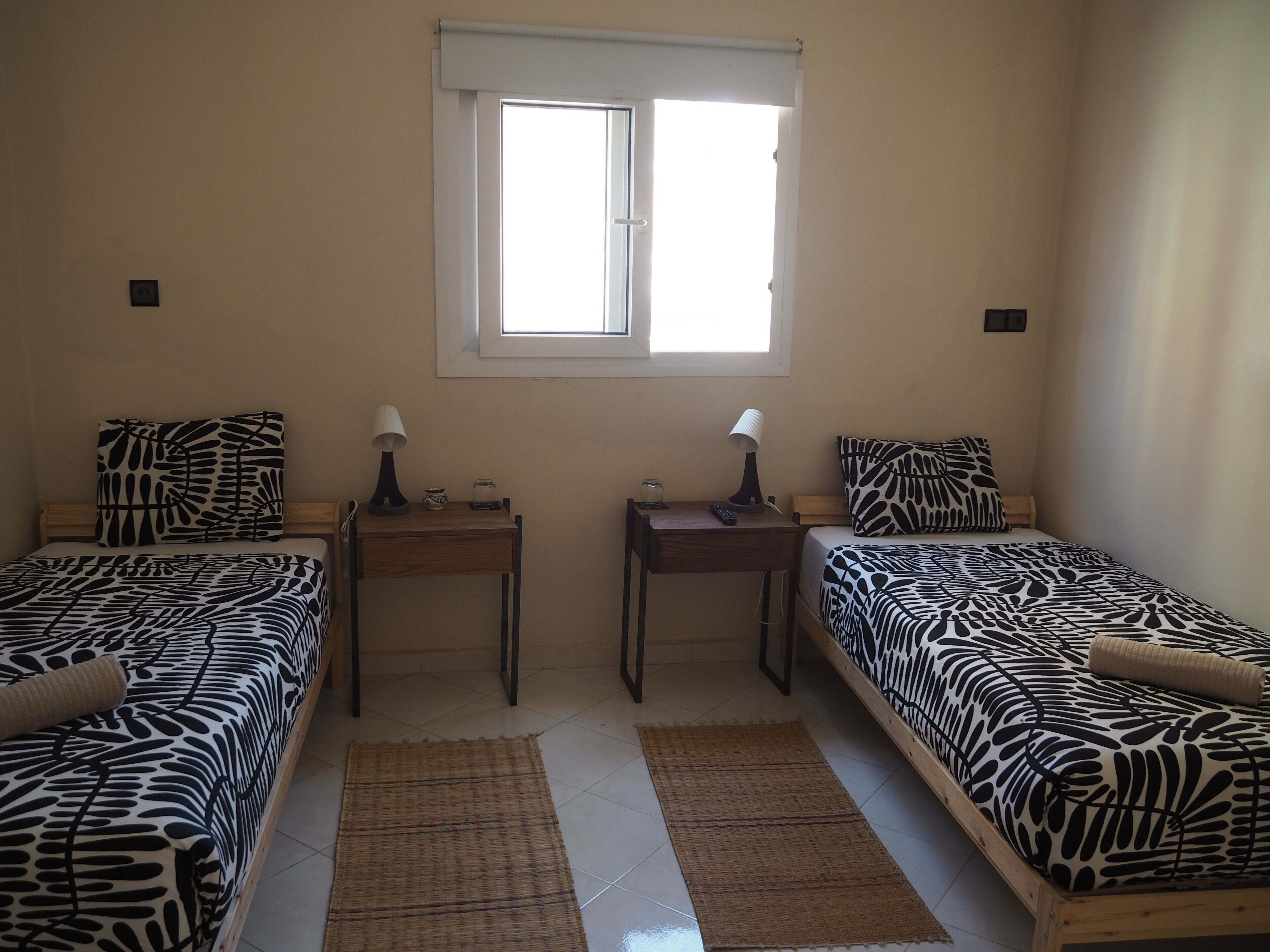 A room with two beds, each with a zebra-patterned bedspread, a small wooden table with a lamp, a window with blinds, and a woven rug on the floor.