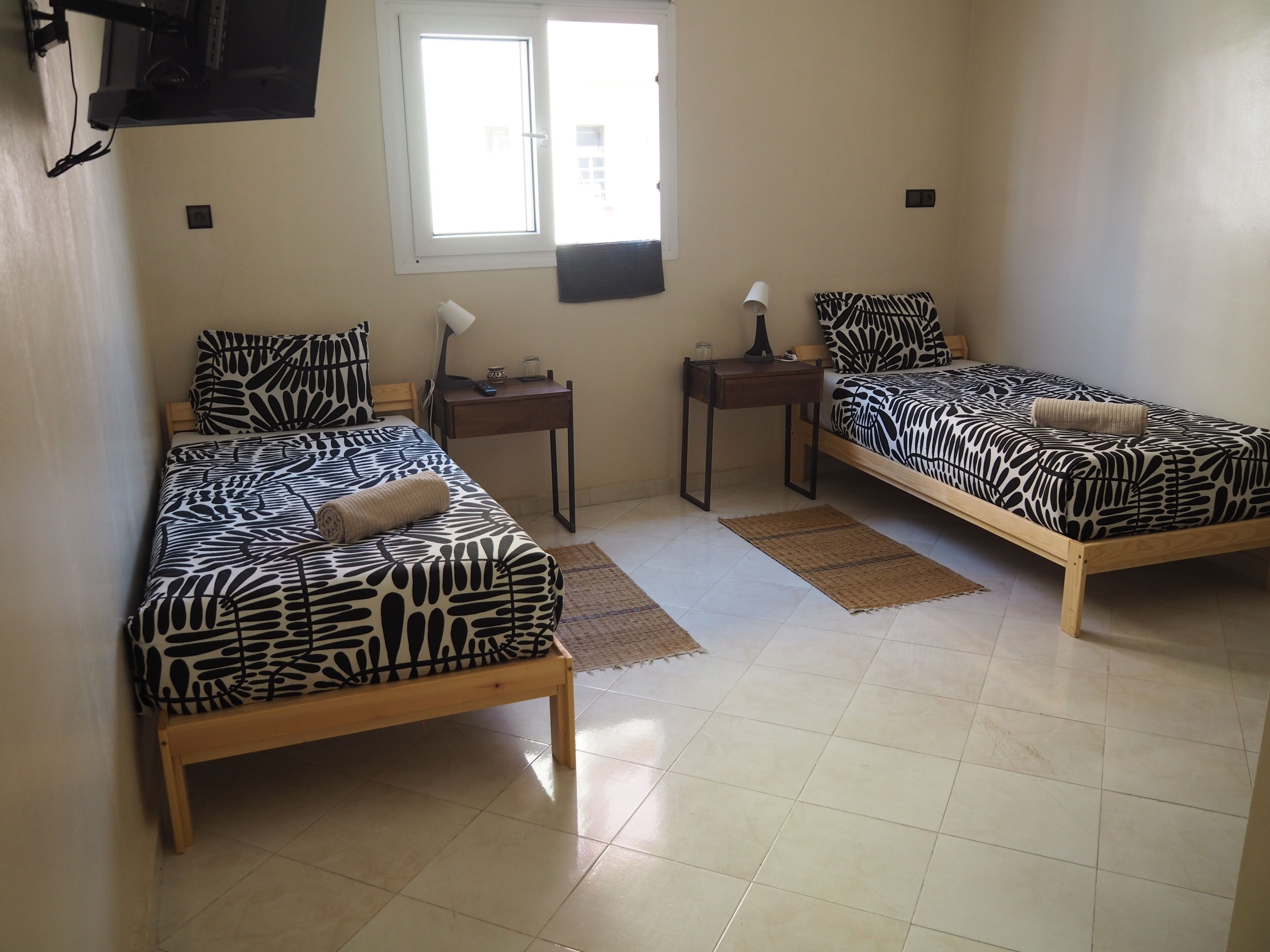 Una habitación con dos camas, cada una con una colcha estampada, una mesita con una lámpara y un ventanal que deja entrar luz natural.