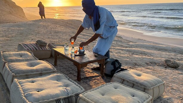 A beach setting with a wooden table and cushions, a person pouring a drink, and a sunset in the background. - Al Mohit Hotel Dakhla (Dakhla)