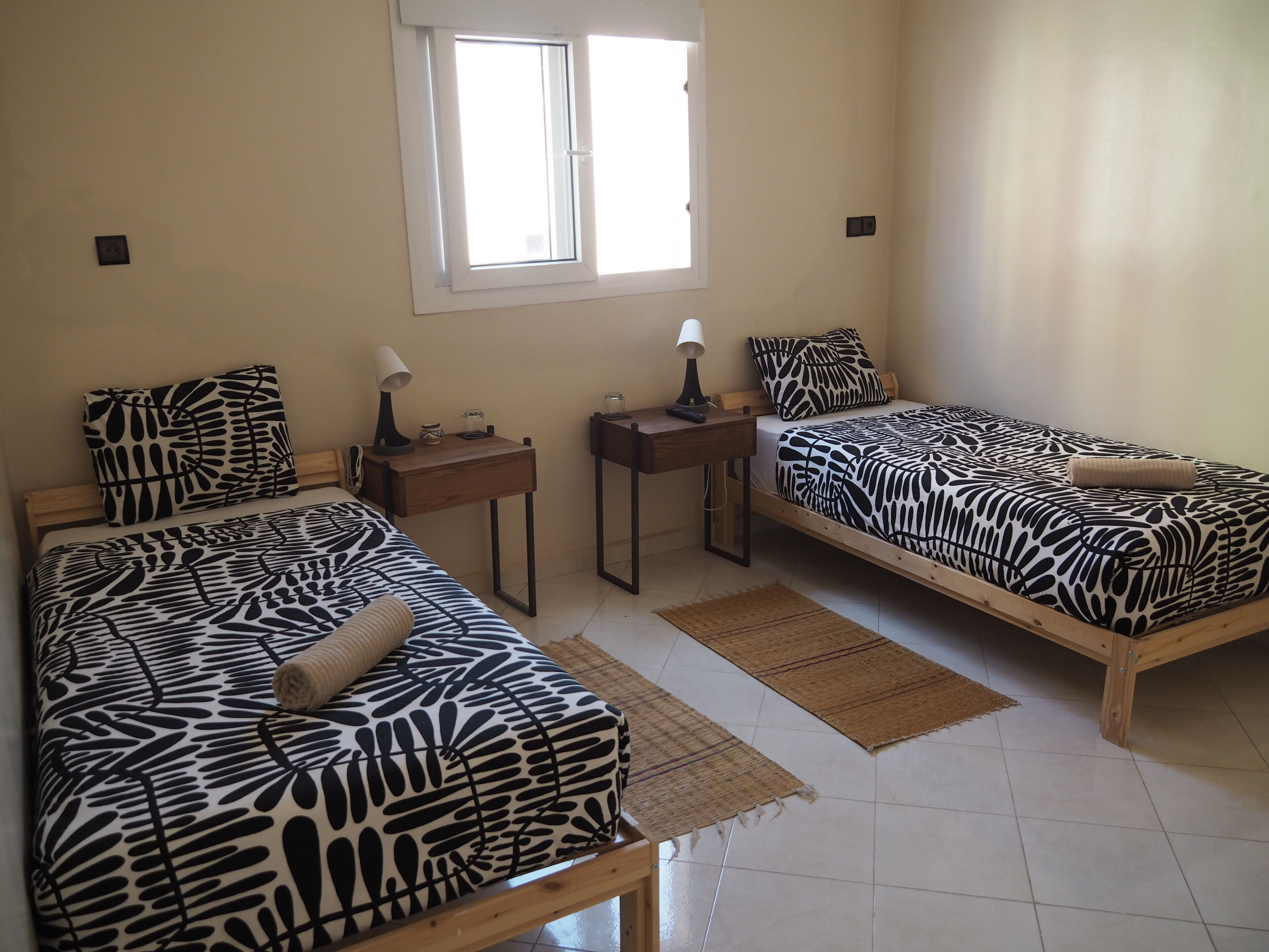 Two single beds with zebra-patterned bedding, a small table with a lamp, a window, and a wall-mounted light switch.