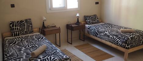 Dos camas individuales con ropa de cama estampada de cebra, una mesita con una lámpara, una ventana y un interruptor de luz en la pared.