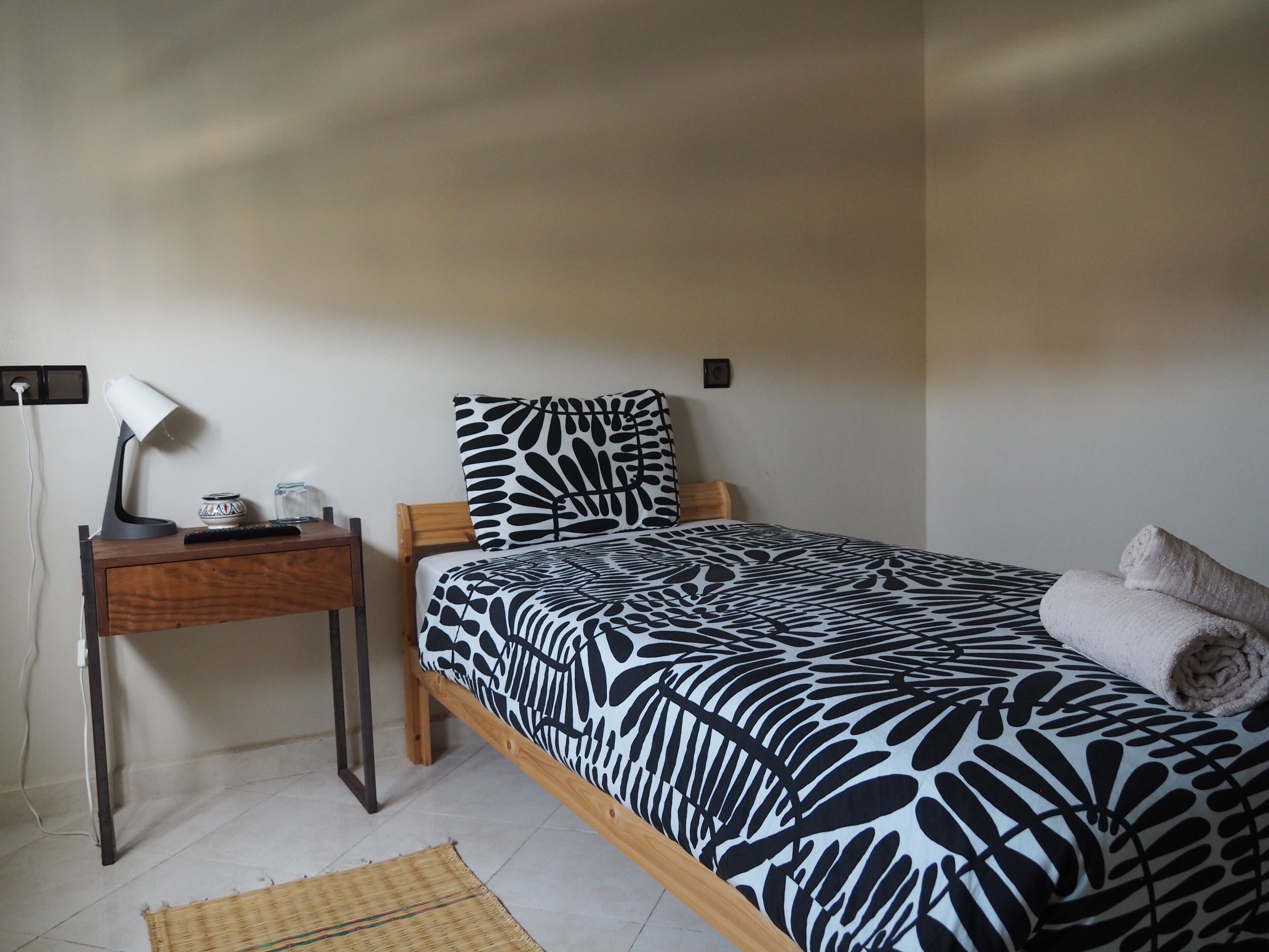 A bedroom with a bed, a nightstand, a lamp, and a wall socket.