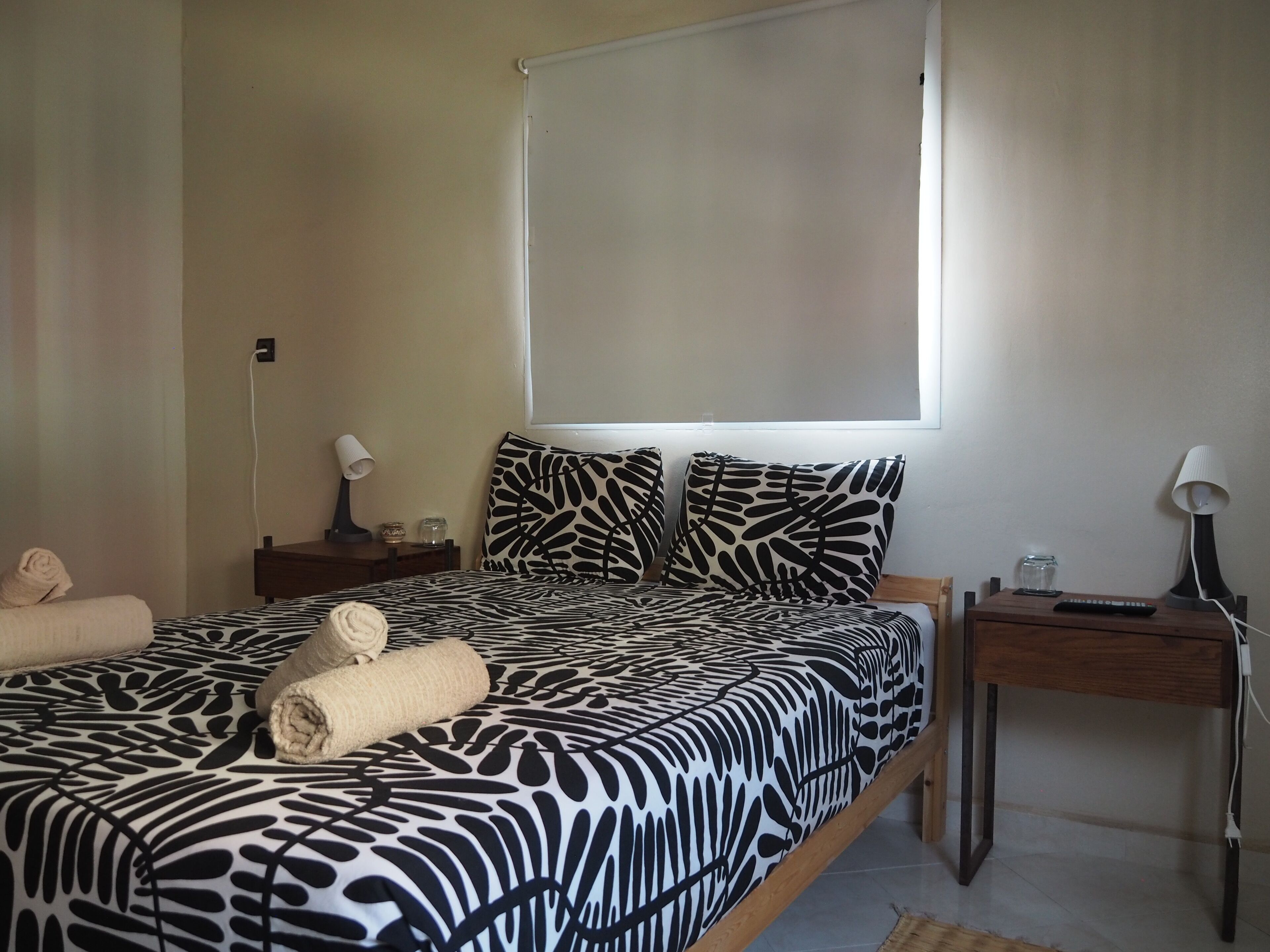 A bedroom with a zebra-patterned bedspread, a large window, a bedside table with a lamp, and a nightstand with a remote control.