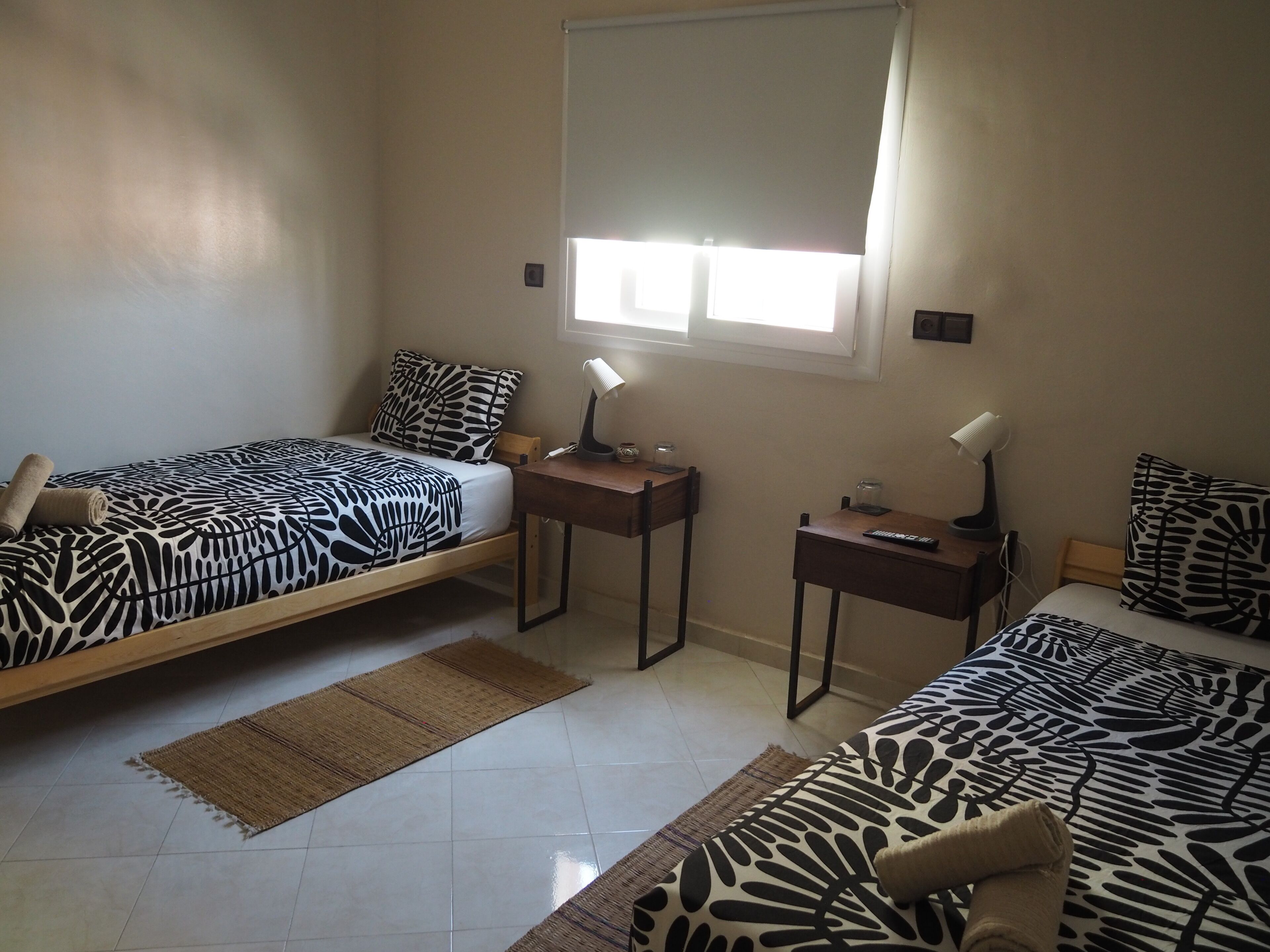 A room with two beds, each with a zebra-patterned bedspread, a small desk with a lamp, and a window with blinds.