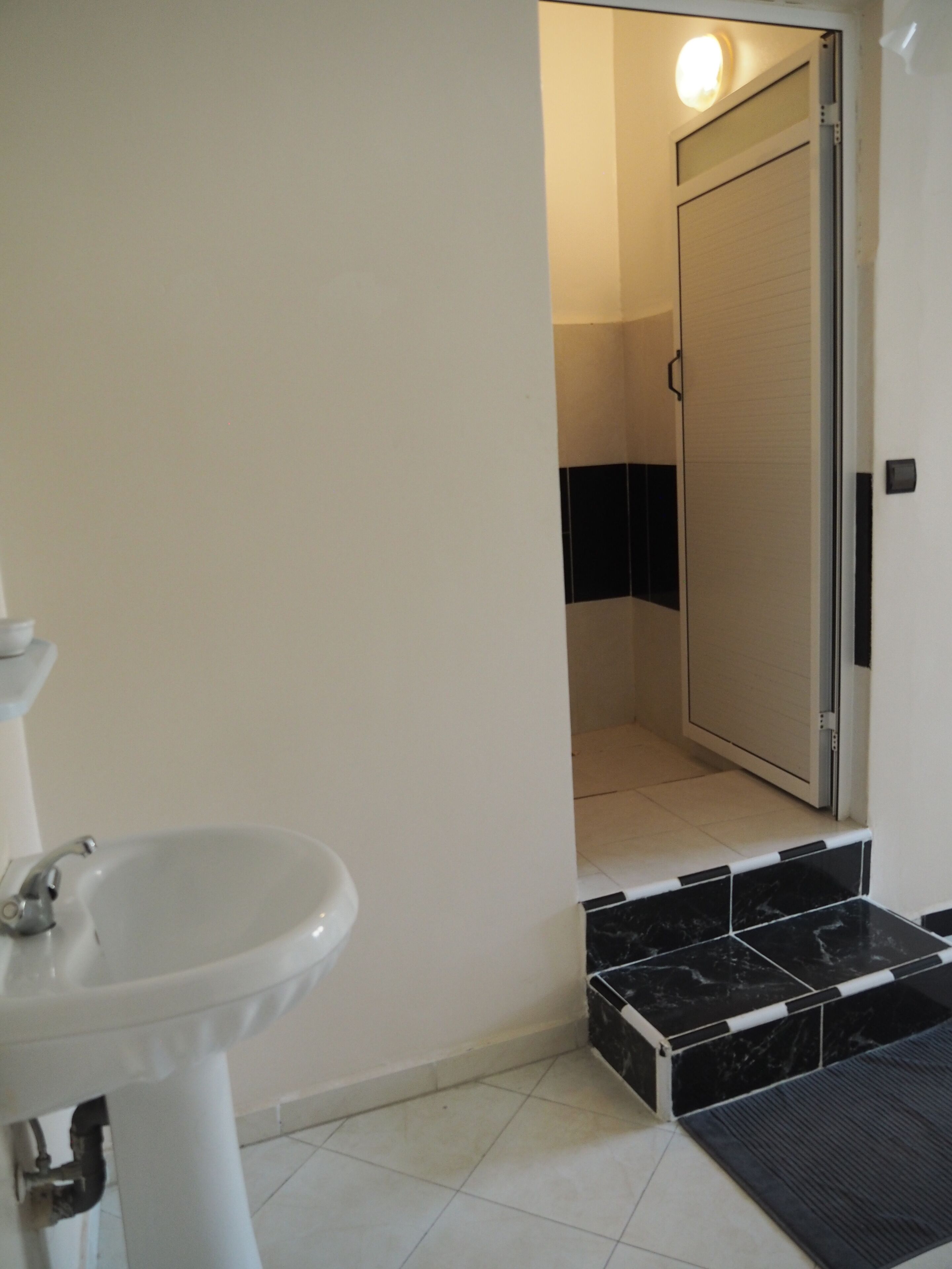 Une salle de bain avec un lavabo blanc, un espace douche et une porte donnant sur une autre pièce.