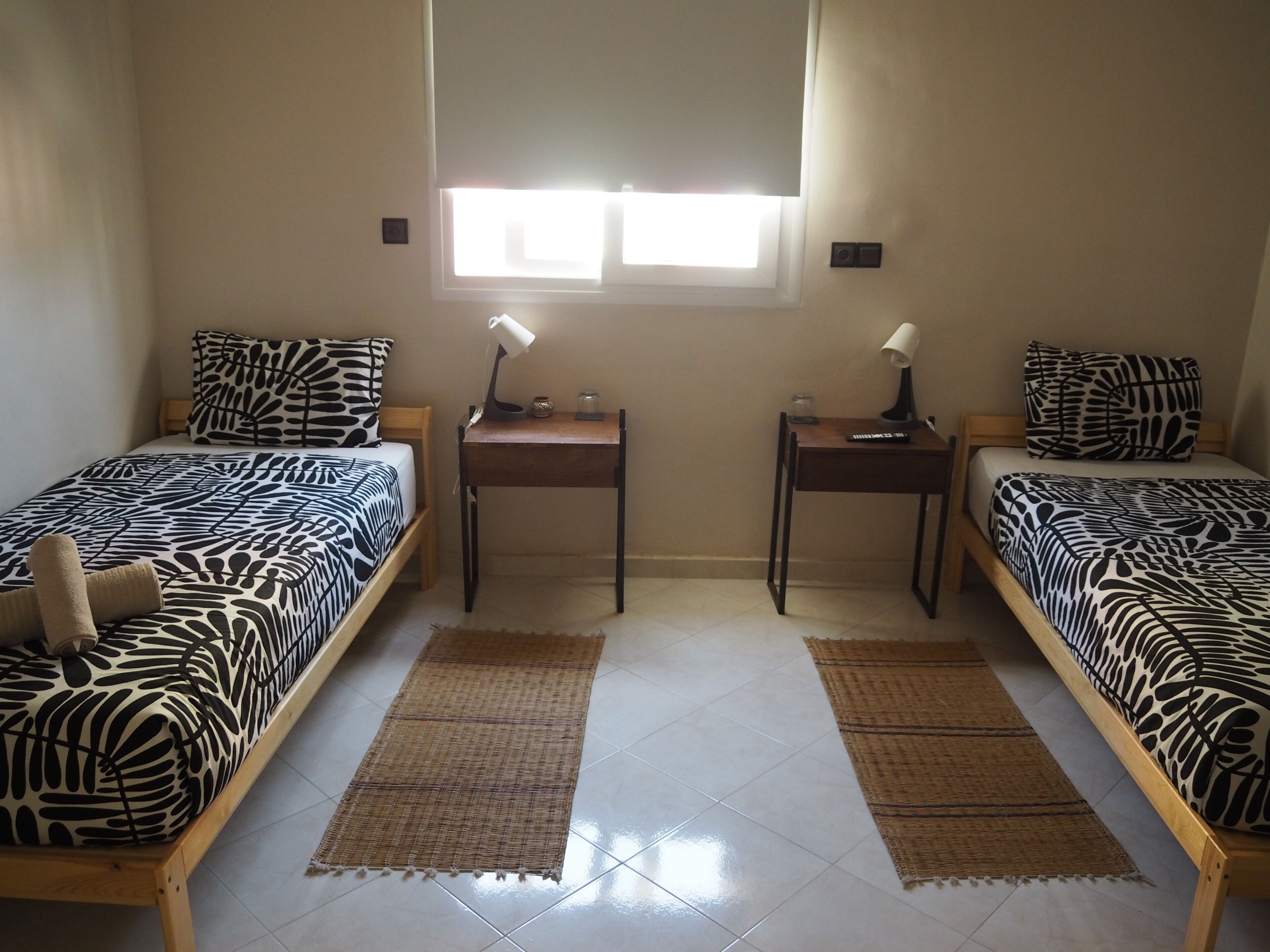 A bedroom with two beds, each with a zebra-patterned bedspread, a small wooden table with a lamp, and a window with blinds.