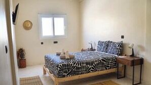 A bedroom with a bed, a bedside table, a lamp, a window, and a wall-mounted hat. - Al Mohit Hotel Dakhla (Dakhla)