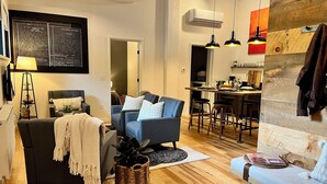 A modern living room with a grey sofa, a blue armchair, and a small dining area with a wooden table and bar stools.