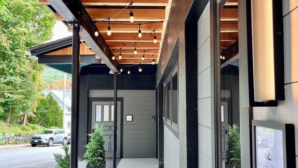 A building entrance with a wooden ceiling and hanging lights, a concrete pathway, and potted plants.