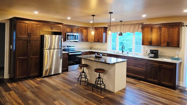 A modern kitchen with wooden cabinets, stainless steel appliances, a central island with stools, and pendant lighting. - Just Like Home, brand new 2 bedroom rancher with full kitchen. (Mohnton)
