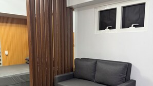 A modern living room with a dark grey sofa, a patterned rug, wooden slats, and a window with blinds. - Studio Unit with Balcony in Manila (Manila)