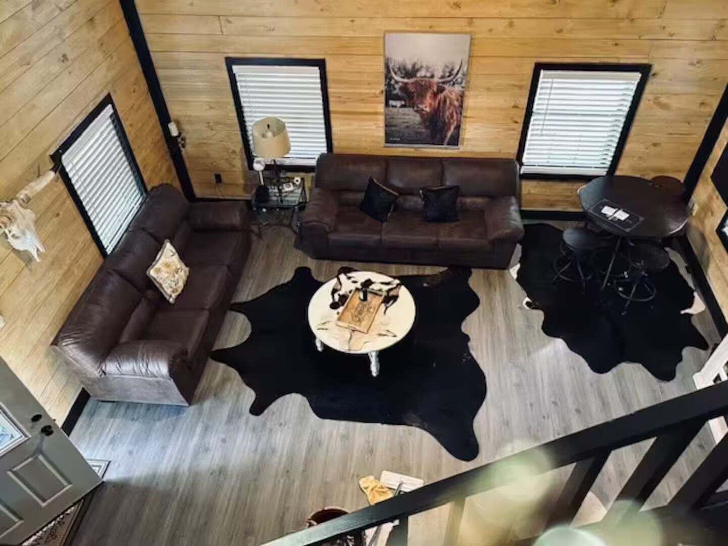 Une pièce aux murs lambrissés, avec un canapé en cuir marron, un tapis en peau de vache noir et blanc, une petite table ronde et un tableau encadré accroché au mur.
