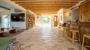 A spacious living area with exposed wooden beams, a stone-tiled floor, a wooden bench, a TV mounted on the wall, and a kitchen area with stools. - Comfortable country house with heated swimming pool in Eure et Loire (Tillay-le-Péneux)