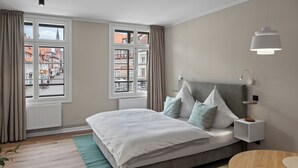 Une chambre moderne avec un grand lit, une table de chevet, une commode et une vue sur la ville grâce à de grandes fenêtres.