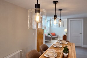 A dining area with a wooden table set for two, clear glass pendant lights, and a view into a living room.